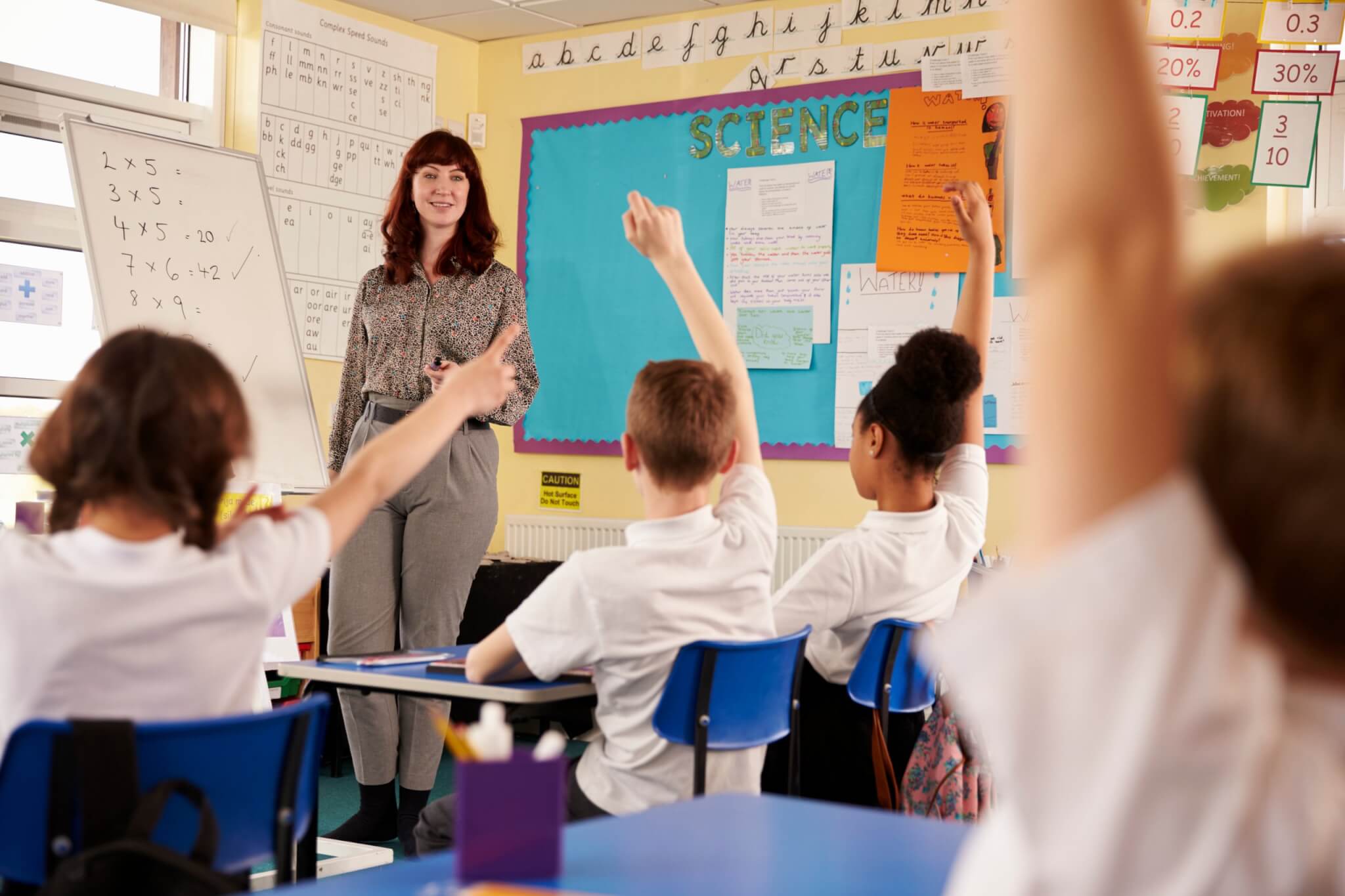 A teacher at the front of a primary classroom with pupils raising their hands