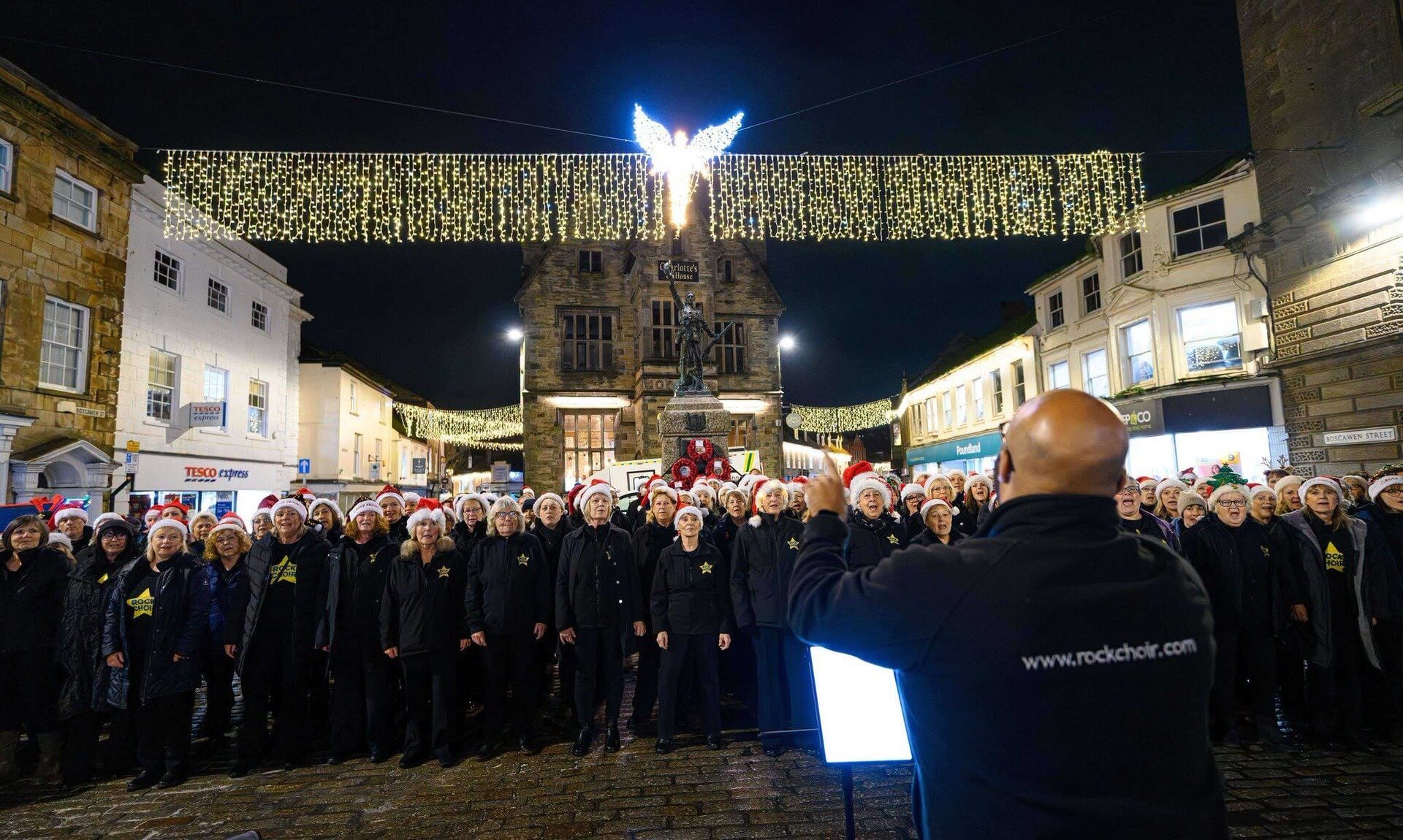 Choir in Santa hats singing beneath Christmas lights on Boscawen Street, Truro