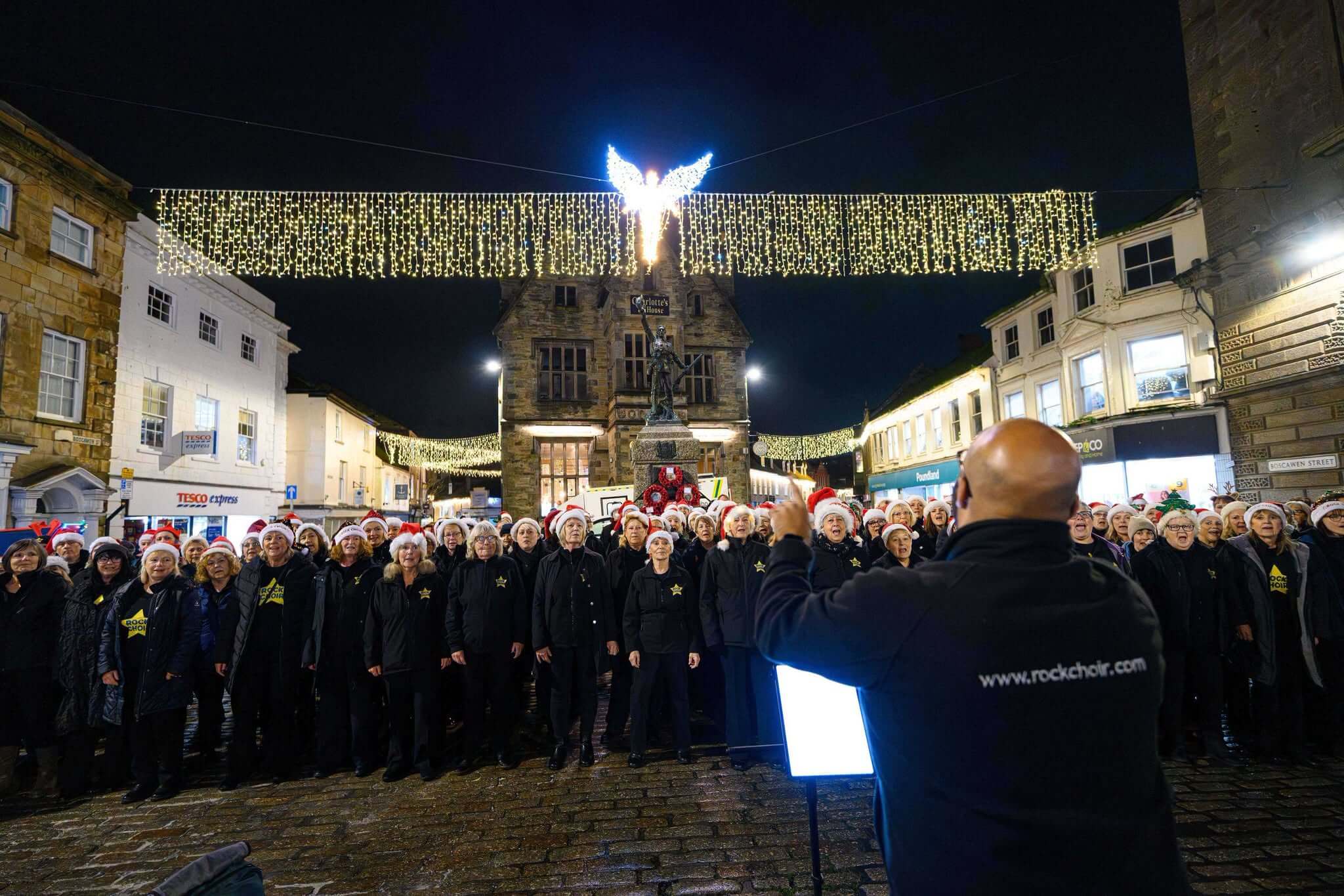 Choir in Santa hats singing beneath Christmas lights on Boscawen Street, Truro