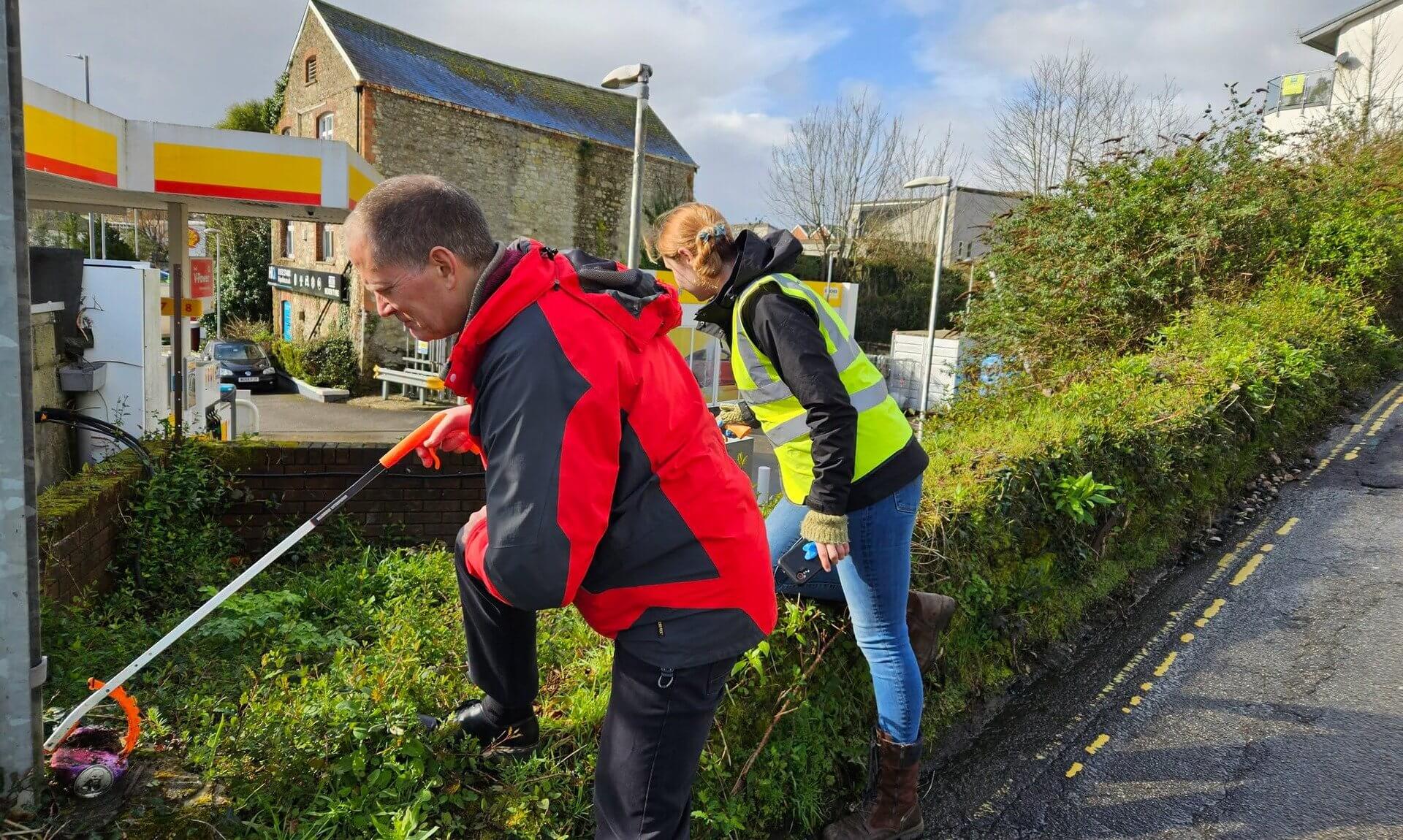 Two volunteers litter picking on a grass verge beside a petrol station