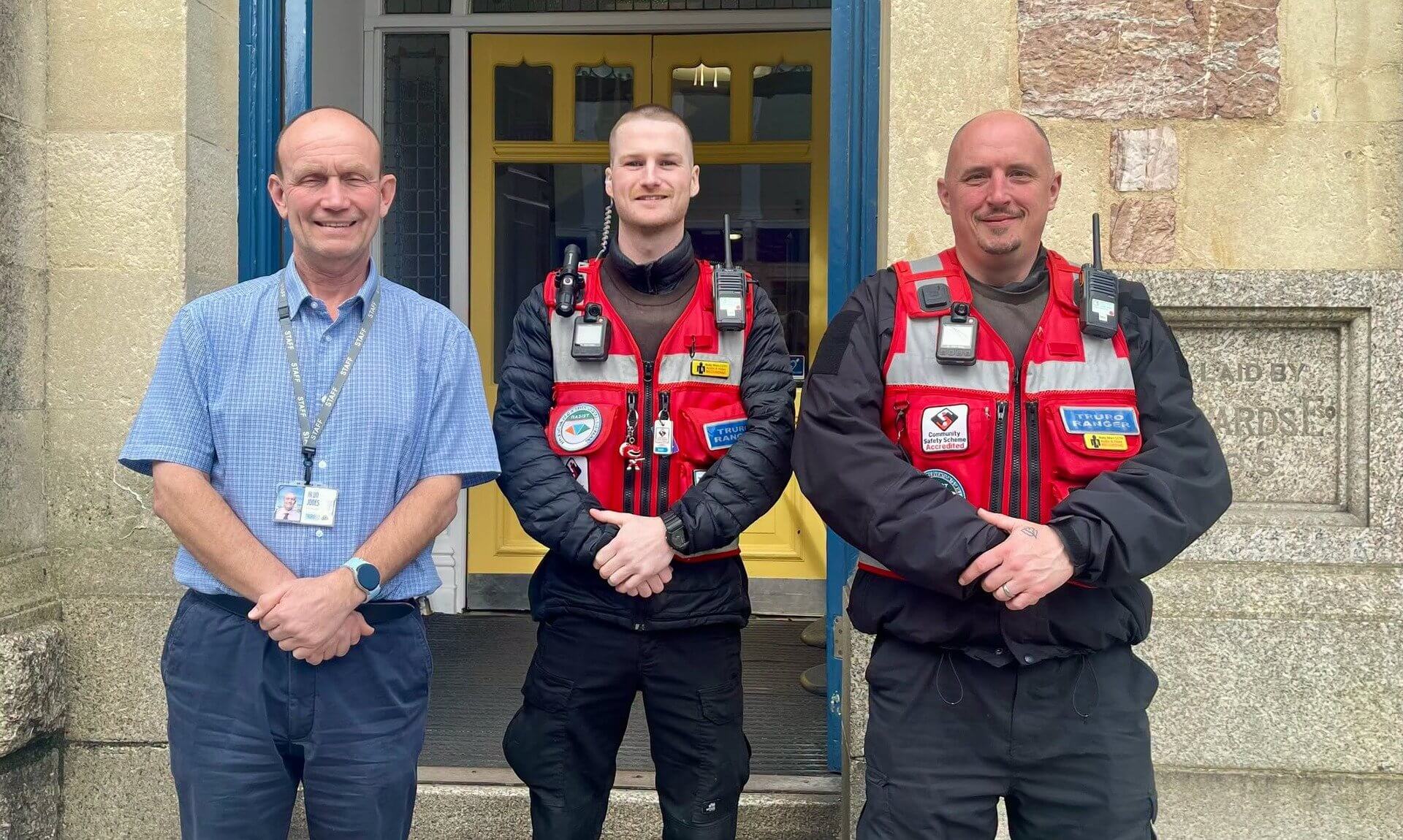 Two Truro Rangers in red patrol vests standing beside a man in a blue shirt outside a building in Truro
