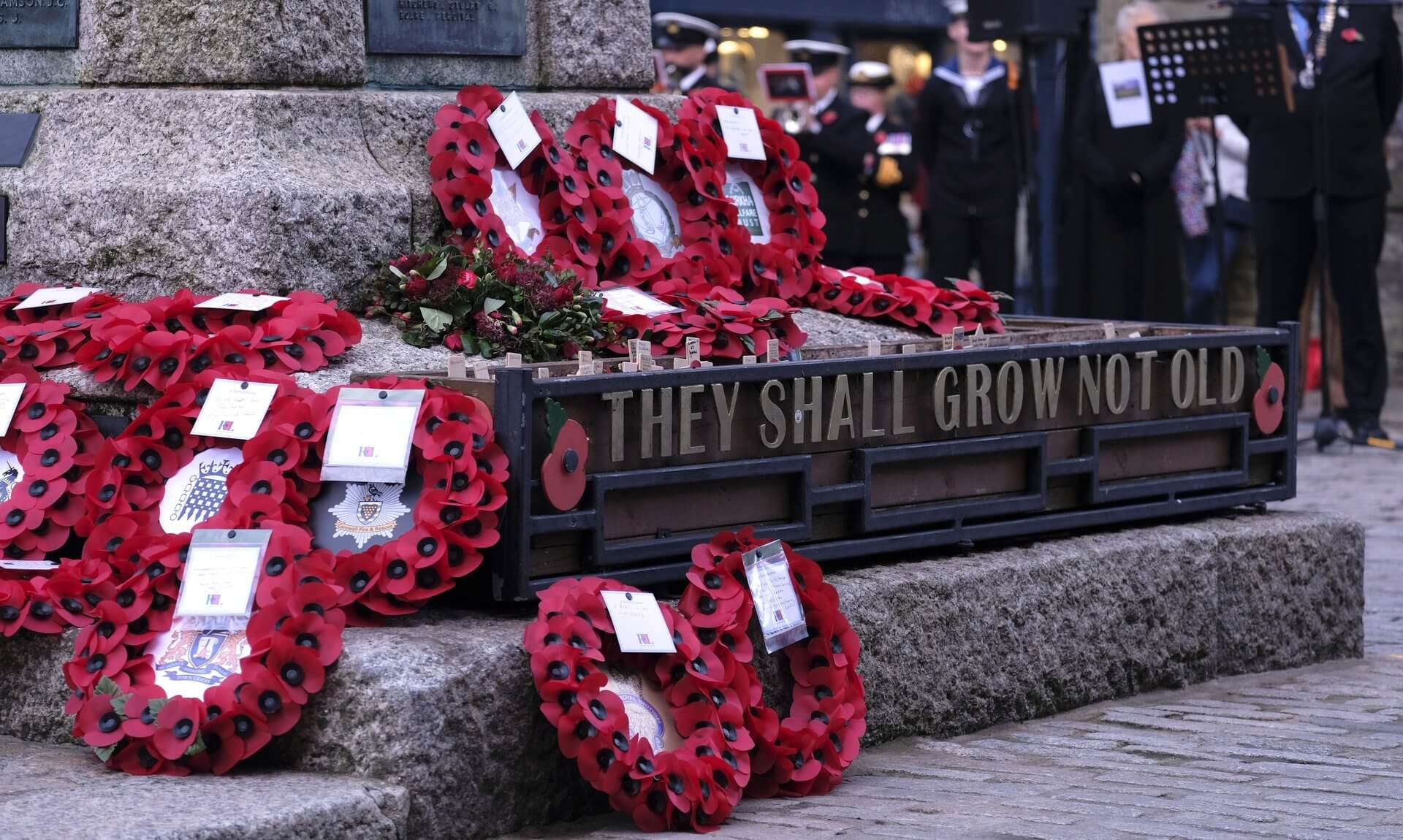 Poppy wreaths laid at the Truro War Memorial beside a planter reading They Shall Grow Not Old