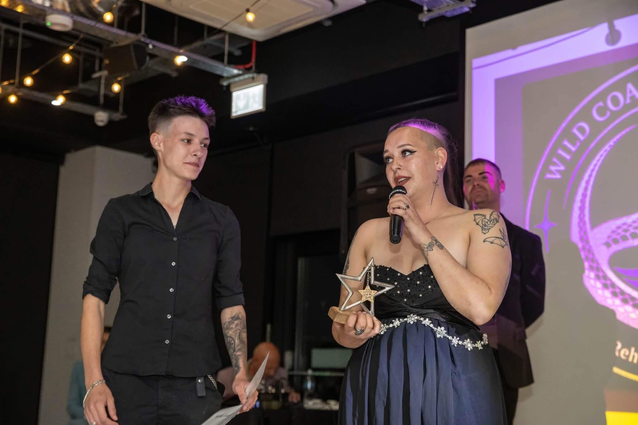 Two people on stage, one speaking into a microphone and holding a star trophy