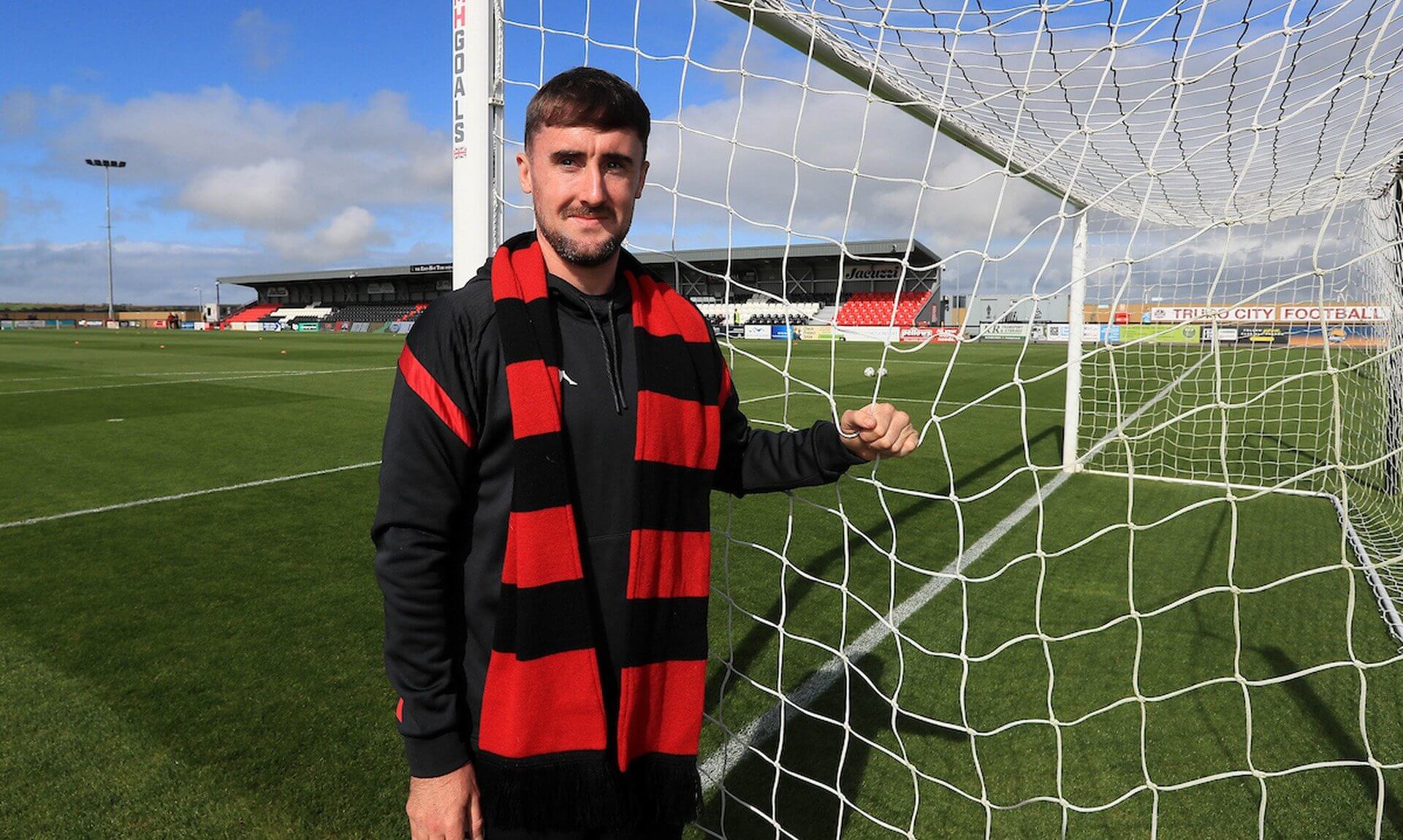 Will Dean in a red and black Truro City scarf beside a goal at Truro City Stadium