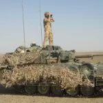 A Scimitar Mk II light tank draped in camouflage netting with a soldier standing on top looking through binoculars