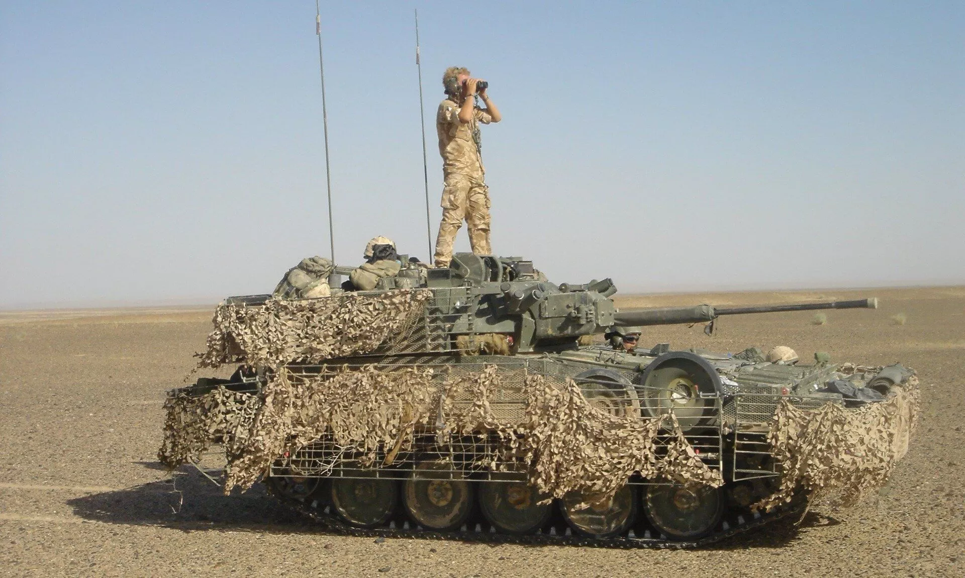 A Scimitar Mk II light tank draped in camouflage netting with a soldier standing on top looking through binoculars