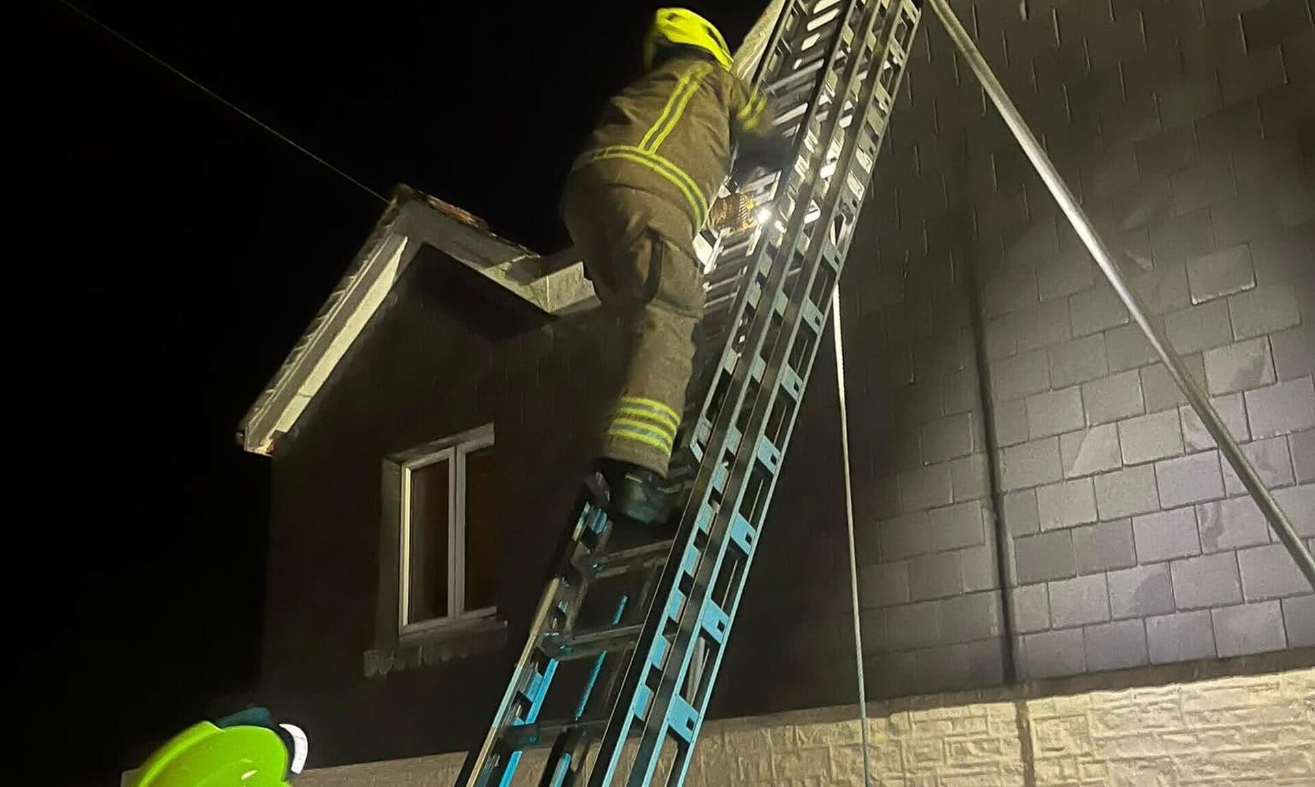 A firefighter climbing a ladder to a chimney at night while a colleague steadies it from below