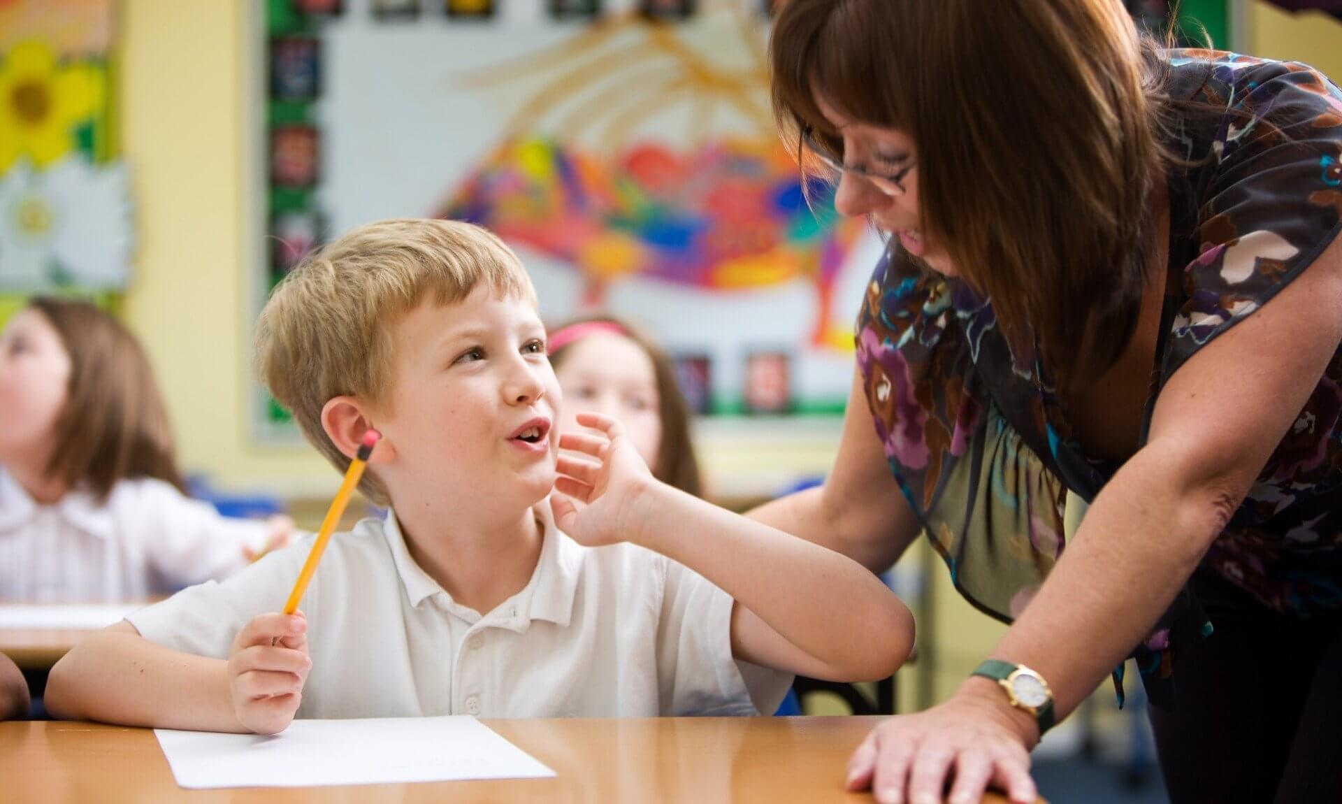 A teacher leaning down to talk with a pupil at a classroom desk