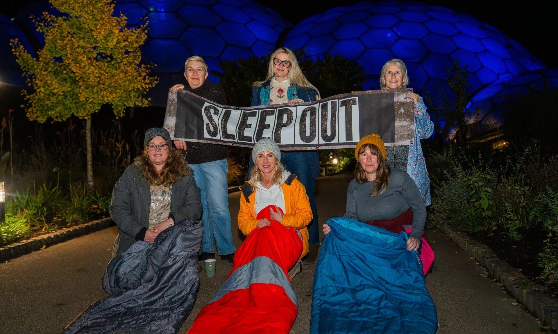 Sleep Out participants in sleeping bags holding a Sleep Out banner in front of the lit-up Eden Project Biomes