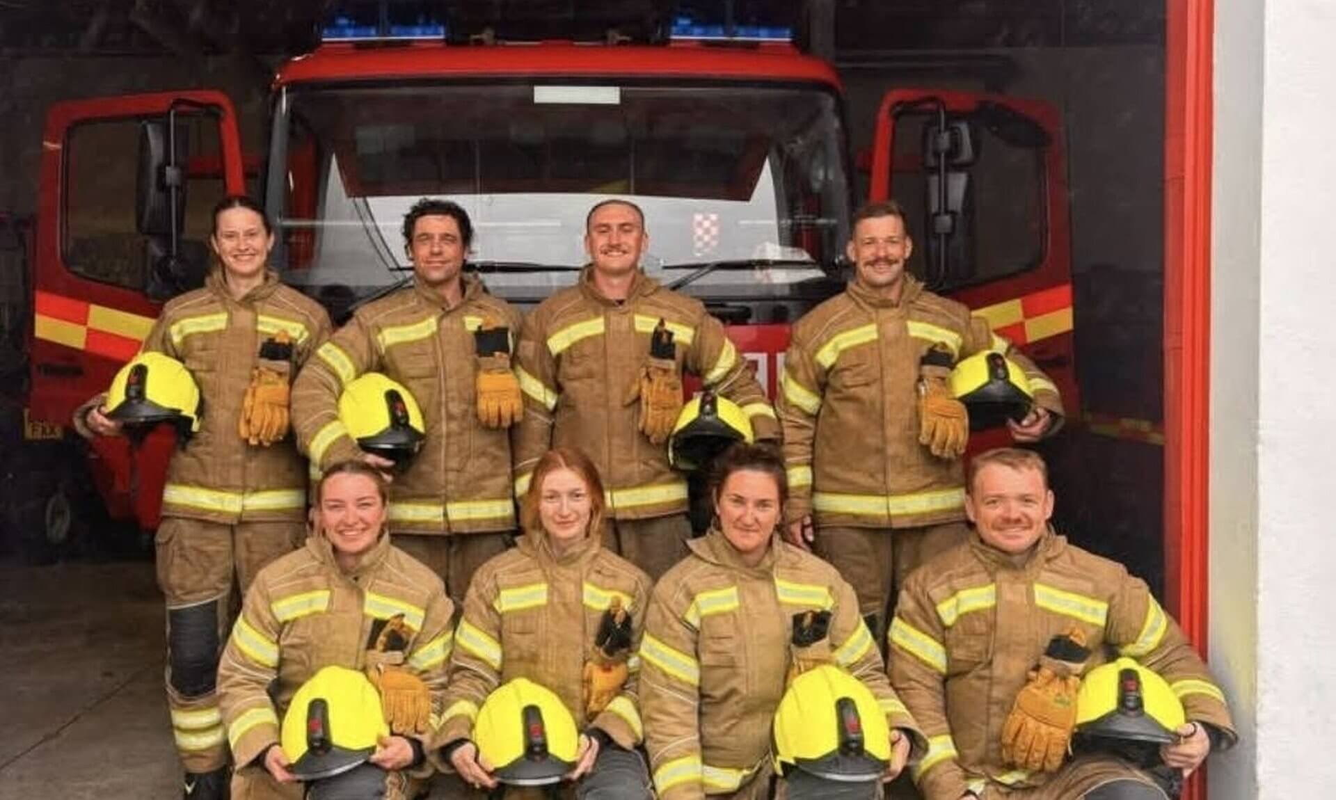 Eight firefighters in kit holding yellow helmets in front of a fire engine at Fowey Community Fire Station