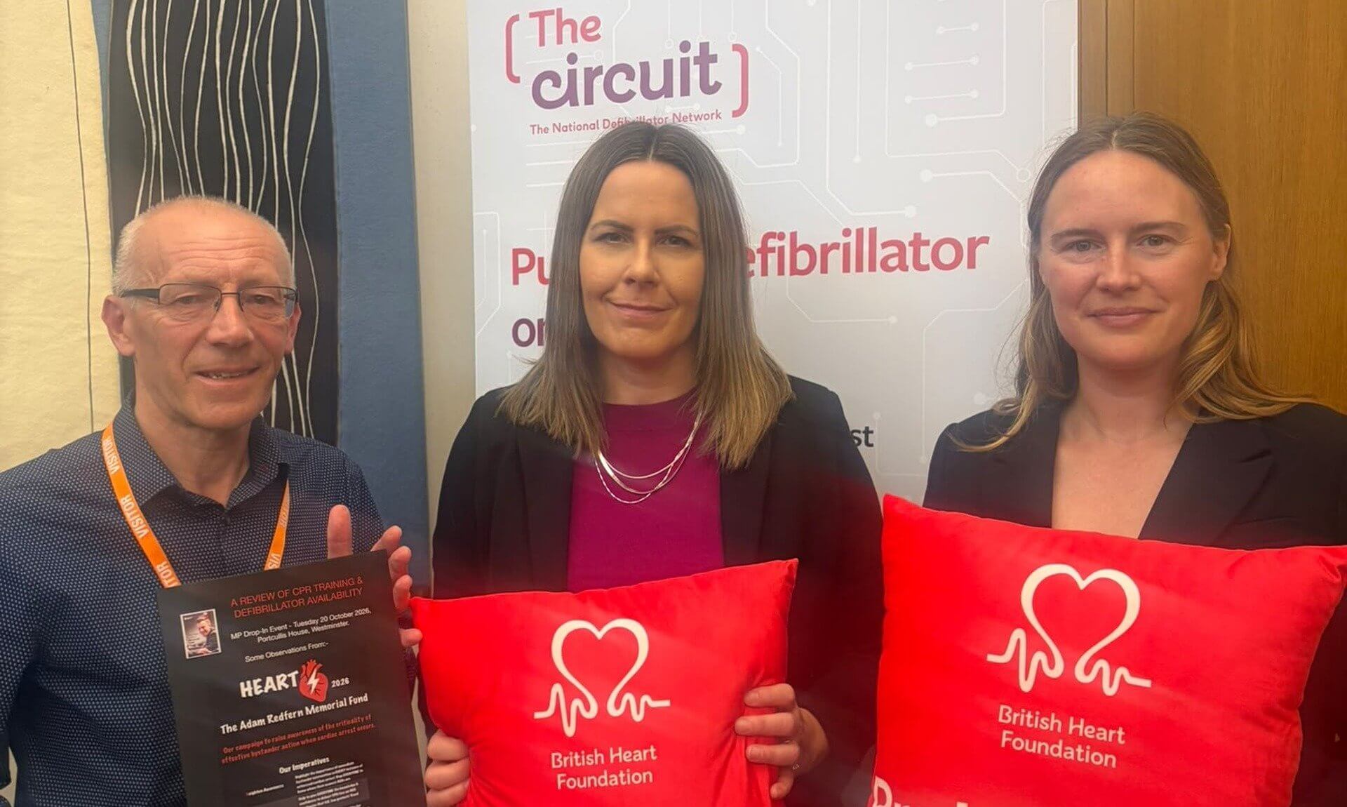 Three people in Parliament holding British Heart Foundation cushions and a defibrillator campaign leaflet