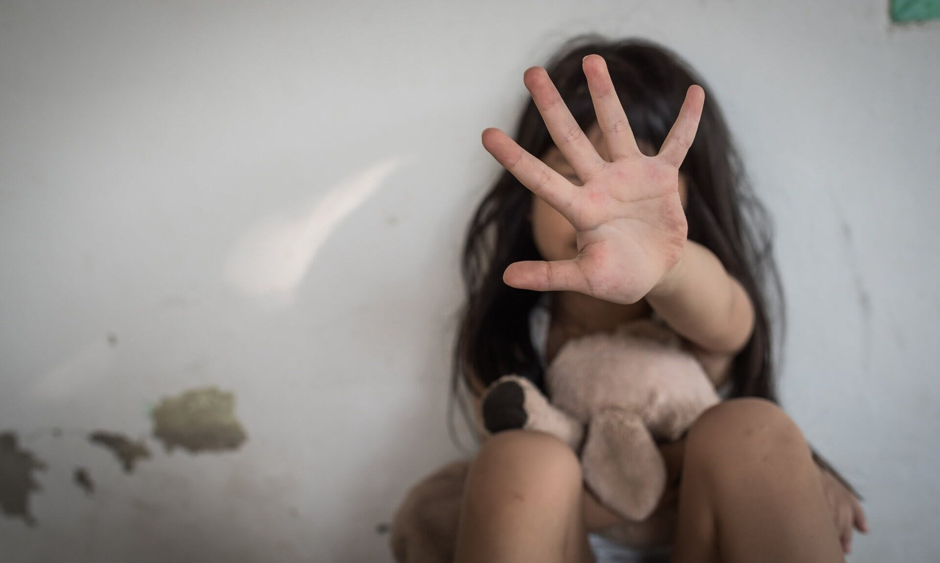 A child sitting against a wall holding a soft toy, with a hand raised towards the camera