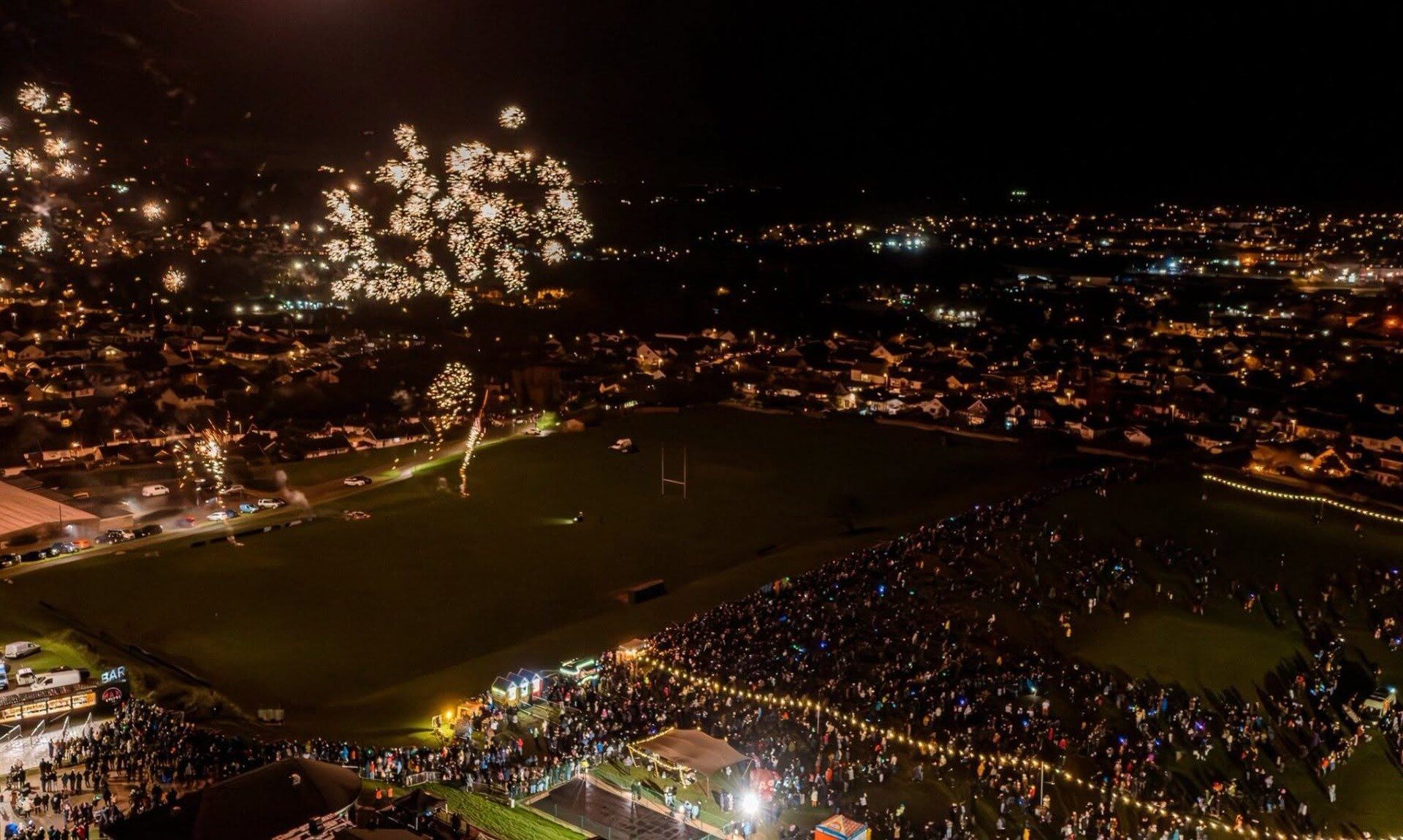 Fireworks over a town at night with crowds gathered on a playing field