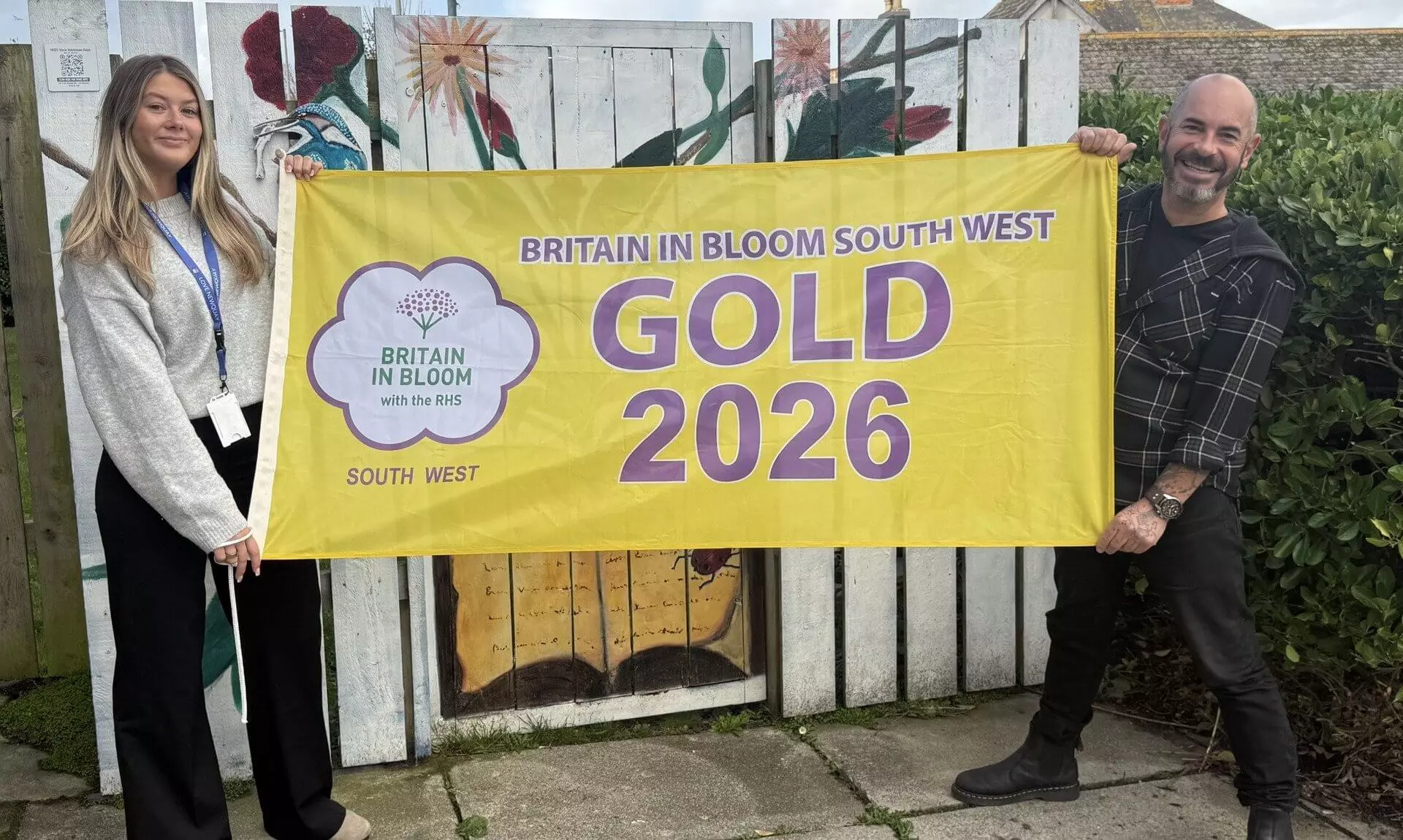 Two people holding a Britain in Bloom South West Gold 2026 flag in front of a painted fence in Newquay