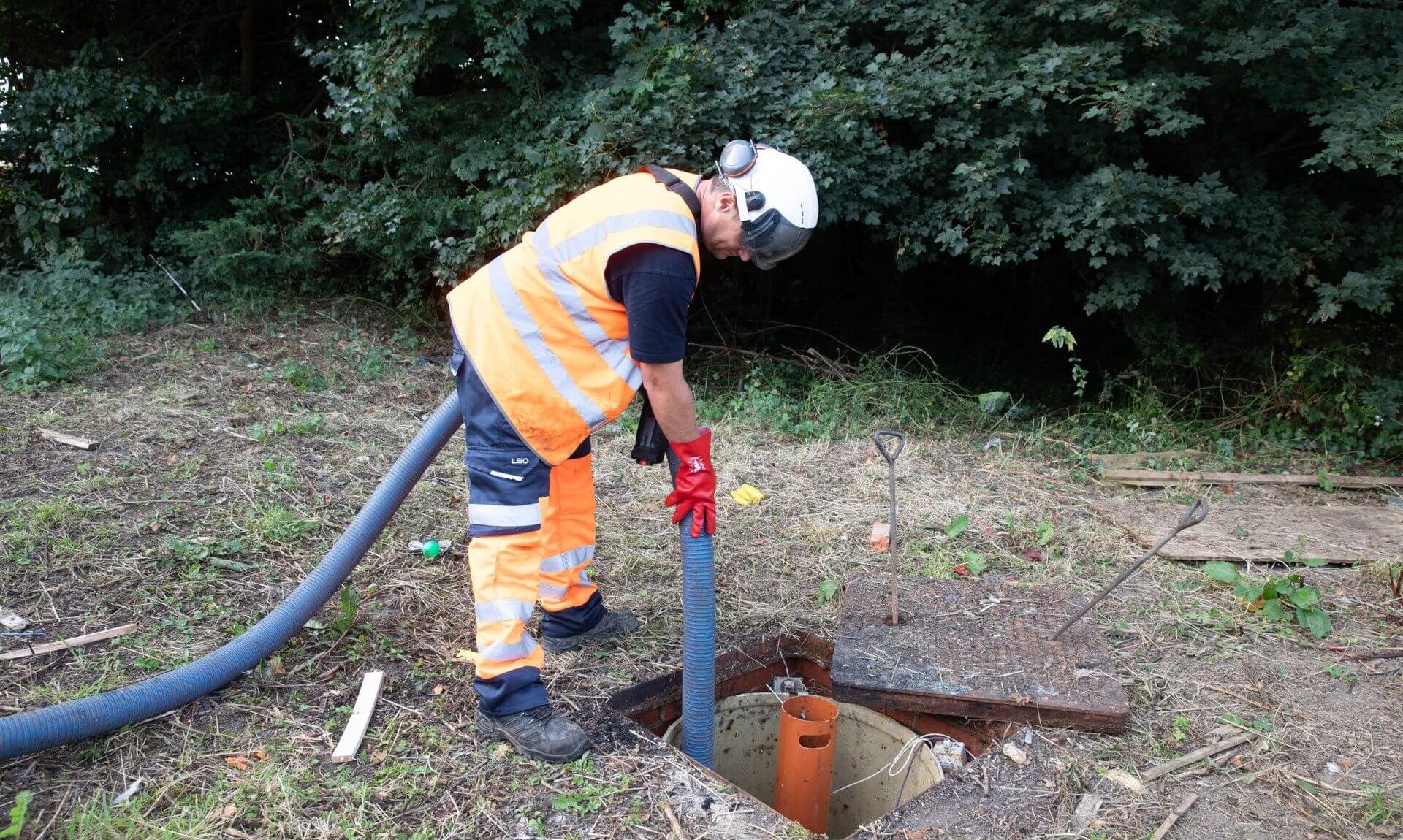 A worker in hi-vis clothing lowers a suction hose into an open septic tank
