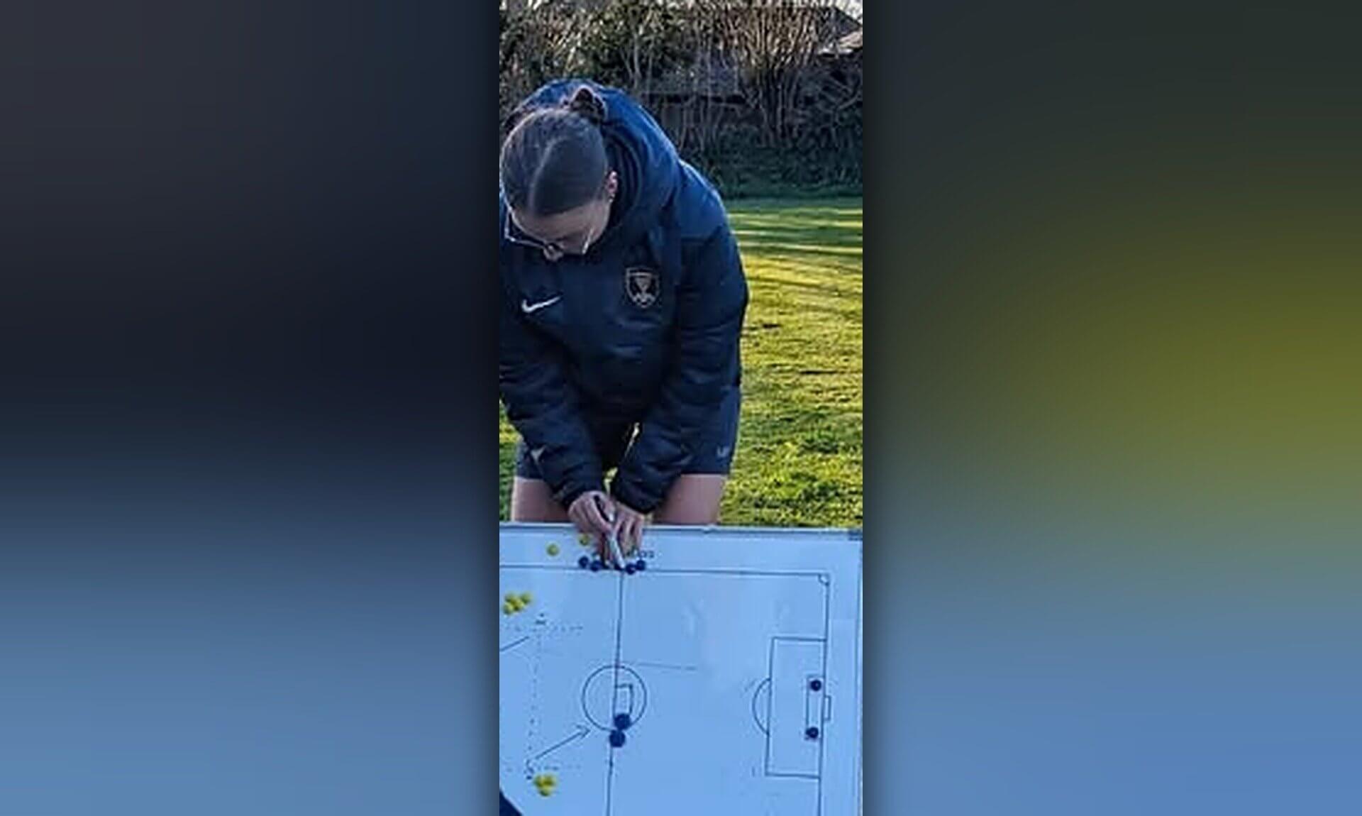 A coach in a navy jacket drawing on a football tactics board on a grass pitch