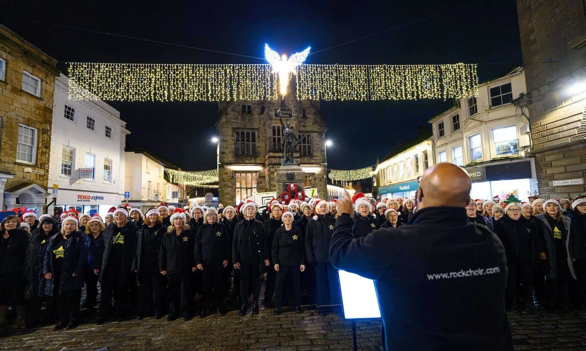 Choir in Santa hats singing beneath Christmas lights on Boscawen Street, Truro
