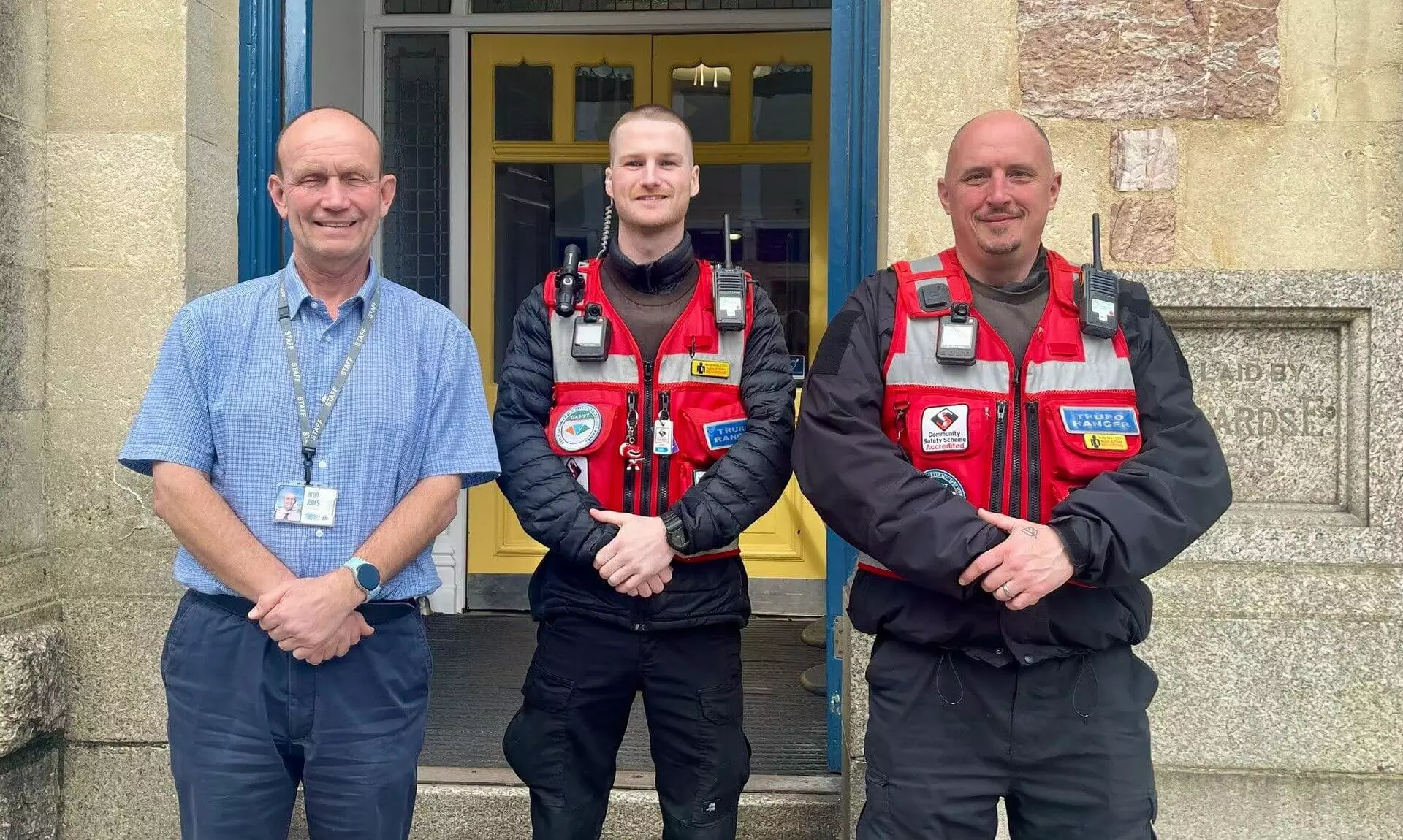Two Truro Rangers in red patrol vests standing beside a man in a blue shirt outside a building in Truro