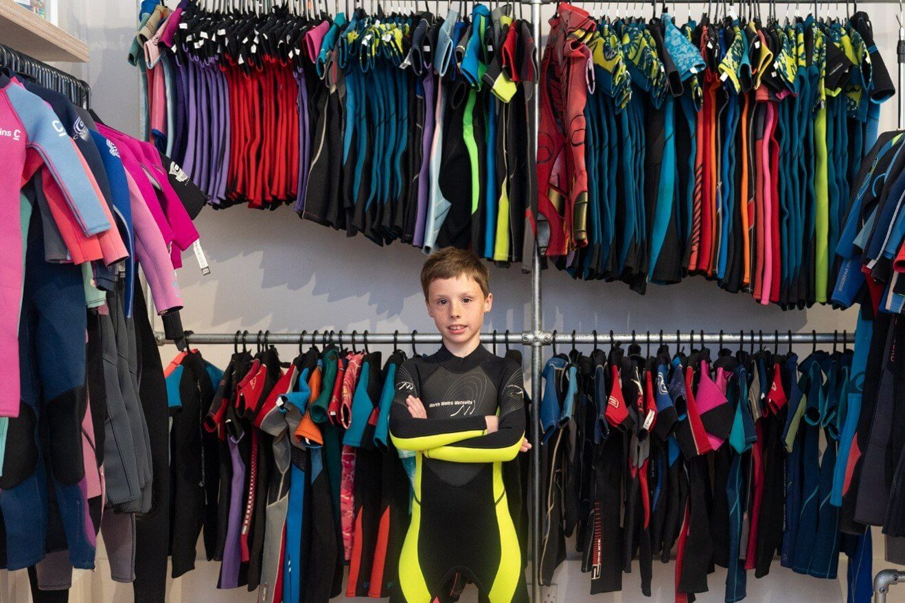 A young boy in a wetsuit standing in front of rails of pre-loved wetsuits