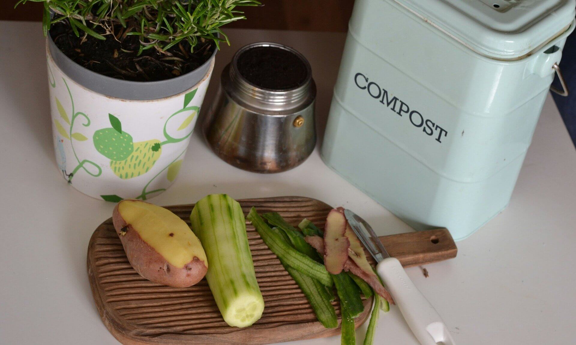 Peeled potato and cucumber with peelings on a chopping board beside a compost caddy